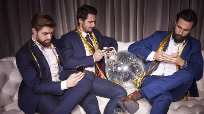 Tre män i stiliga kostymer sitter i soffa och ser på sina telefoner