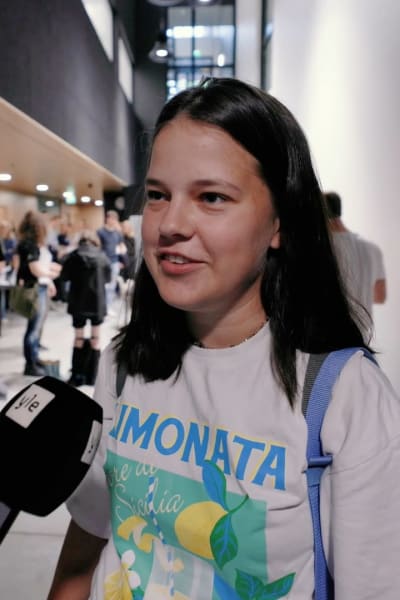 En ung kvinna blir intervjuad med mikrofon. Personen har axellångt mörkt hår och är iklädd en vit t-skjorta med italienskt tryck med texten "Limonata". Personen bär också på en ryggsäck. I bakgrunden syns många personer.