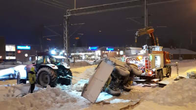 En plogbil har vält över tågräls. En man står bredvid och flera fordon försöker flytta den. Snö på marken.