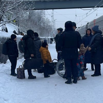 Joukko ihmisiä seisoo lumisella juna-aseman laiturilla. Viereisellä raiteella on pysähtynyt juna.