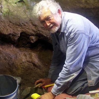 Antropologi Lawrence Straus polvillaan luolassa, kädessä ja vieressä arkeologin välineistöä.
