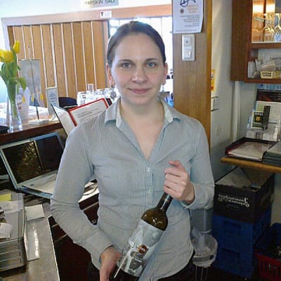 Sommelier Heidi Mäkinen seisoo ravintolan tiskin takana ja pitää viinipulloa käsissään.