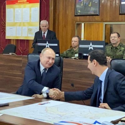 Vladimir Putin ja Bašar al-Assad kättelevät kokoussalissa, jossa istuu myös muita ihmisiä.
