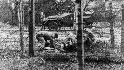 En svartvit bild från 1960-talet. Bilden är ganska grynig men man ser att fyra uniformsklädda män sitter i huk vid något på marken. Mellan dem o fotografen finns taggtråd och i bakgrunden en pansarvagn.