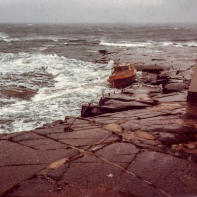 Stormigt hav, liten träbåt förtöjd vid klippa.