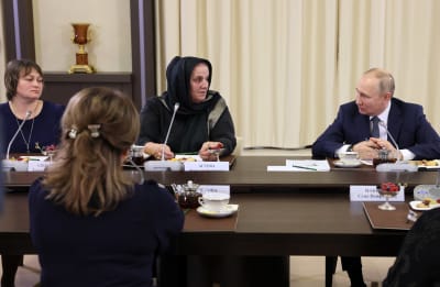Vladimir Putin sitter i ett bord och talar med flera kvinnor. De har neutrala ansiktsuttryck.