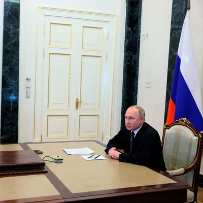 Putin istuu kuvan oikeassa laidassa pöydän päässä kädet ristissä edessään. Takana näkyy Venäjän lippu. Pöydällä on televisioruutu, jossa näkyy turvallisuusneuvoston jäseniä.