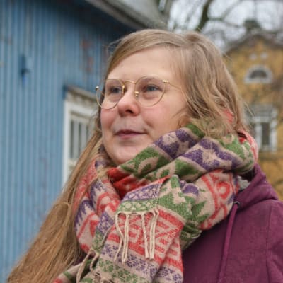 En kvinna i mörkröd jacka och färggrann halsduk står utomhus framför ett blått trähus och ett gult trähus på en gata.