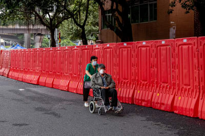 En kvinna skuffar en man i rullstol. De bär båda munskydd. Bakom dem ett staket i röd plast.