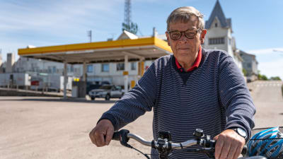 Äldre man leder sin elcykel och ser mot kameran, i bakgrunden en bensinmack i centrum av Hangö.