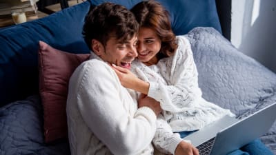 Ett ungt par halvligger i en säng med en dator i famnen och kittlar varandra.