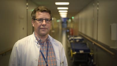 En man med läkarrock ser mot kameran i en sjukhuskorridor