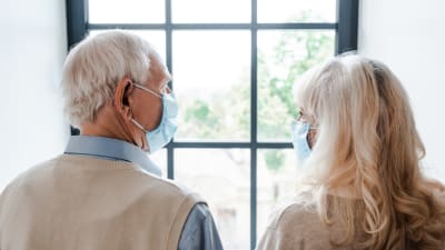 Äldre par med munskydd ser ut genom fönstret. 