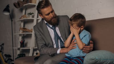 En man i kostym tröstar en liten gråtande pojke, de sitter i en soffa