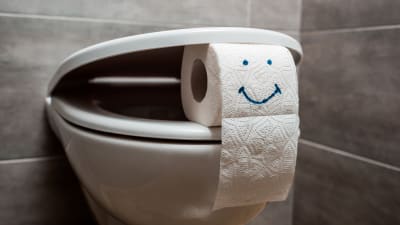 En toalettstol där locket är på glänt med hjälp av en toarulle som har ett leende på sig.