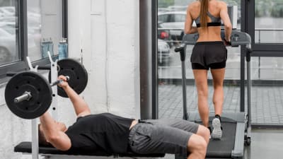 I förgrunden en man som lyfter vikter på gym, i bakgrunden en kvinna som springer på löpmatta.