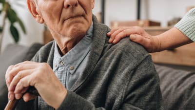En äldre man håller i en käpp. En kvinna håller i honom på axeln medan han tittar bakåt.