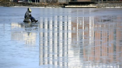 En pilkare sitter på den blanka isen i dagsljus utanför academill och silorna. Man ser reflektionen av silorna i isen.