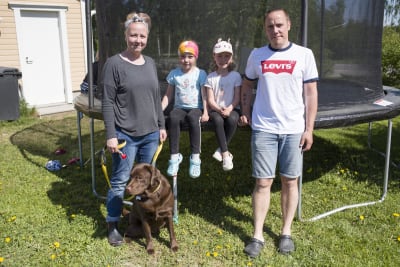 En kvinna, två barn (flickor) och en man står framför en stor studsmatta. Framför dem sitter en brun labrador. Det är sommar och solen skiner.