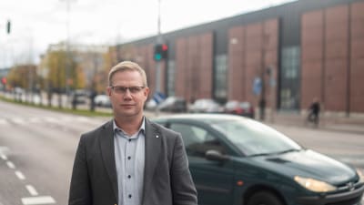 Direktören för den svenska servicehelheten vid sektorn för fostran och utbildning, Niclas Grönholm, står ute framför en väg. Det är höst. En bil kör förbi, Grönholm ser allvarsam ut och tittar åt höger.