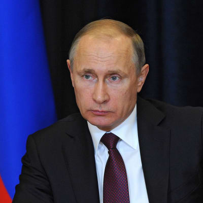 Putin katsoo vakavana kuvan vasenta reunaa kohti. Hänellä on tumma puku, tummanpunainen kravatti, jossa on sinistä kuviota. Taustalla näkyy Venäjän lippu.