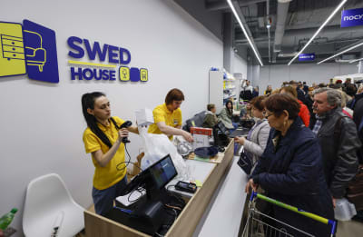 Två butiksbiträden i en kassa medan människor köar. På väggen står det "Swed house".