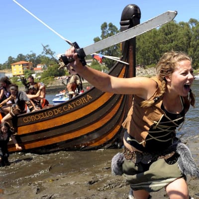 Kuvitteelliseen viikinkiasuun pukeutunut nainen ryntää miekkaa heiluttaen veneestä rantaan.  