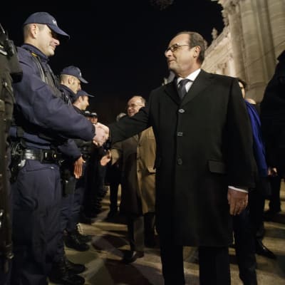 Ranskan presidentti François Hollande tarkastamassa turvallisuusjoukkojen riviä illalla. Hollande kättelee poliisia, joka on sinisessä univormussa, lippalakki päässään. Hollandella on musta päällystakki, musta kravatti. Kuvassa vasemmalla etualalla on toisen poliisin konepistooli, joka roikkuu varustevyöstä.