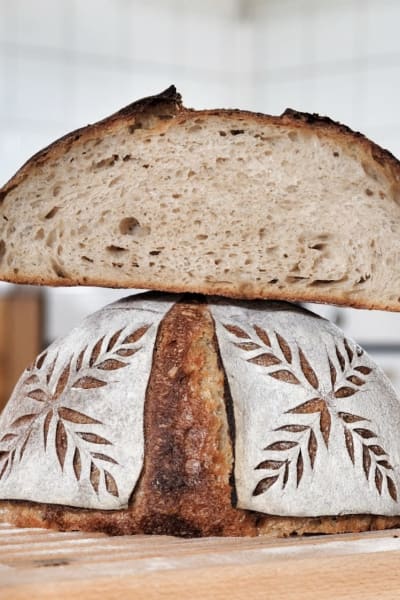 Ett surdegsbröd med dekorerad skorpa. Ovanpå brödet ligger ett uppskuret surdegsbröd.