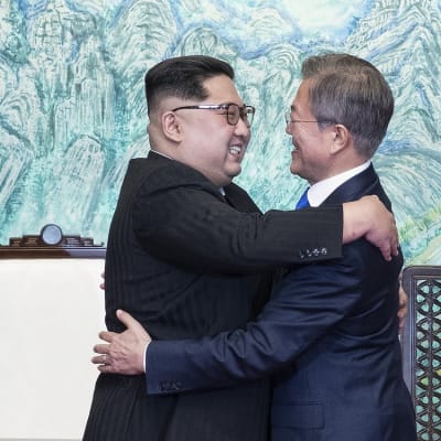 Kim ja Moon halaavat hymyillen kokoushuoneessa. Kimillä on musta maolaishenkinen puku ja Moonilla tummansininen puku.