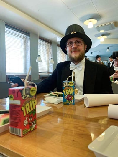 Tom Pakkanen sitter vid ett bord med två små safter framför sig. Han är iklädd i kostym och en hög svart hatt och ser mot kameran.