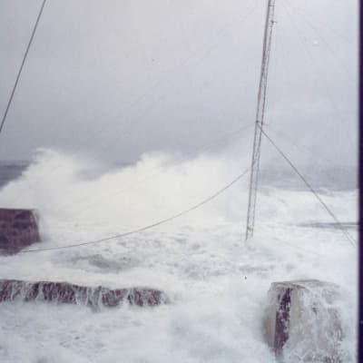 Stormigt hav som sköljer upp över klippor, mast som böjer sig för vinden.