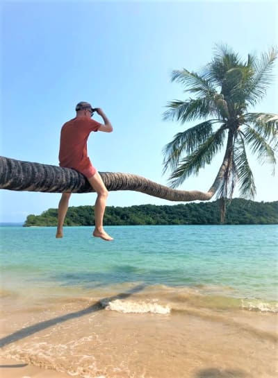 Mies istuu hajareisin poikittain meren päällä kasvavan palmun rungolla.