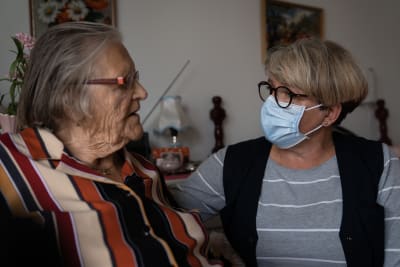 Närvårdaren Manuela Nordström, klädd i grått med svart väst och munskydd, talar med Gretel Gestrin, i randig blus