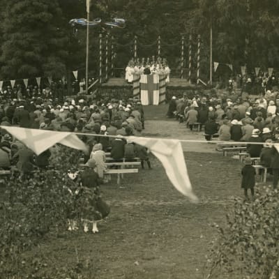Publik på bänkar utomhus som ser på en scen i trä som utsmyckats med Finlands flagga och lövgirlanger. Bilden är svartvit och över hundra år gammal.