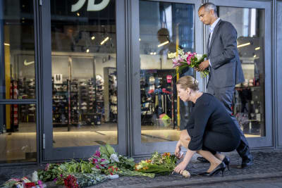 Mette Frederiksen och  Mattias Tesfaye lägger ner blommor utanför en butiksdörr. Det ligger fler buketter på stenläggningen.