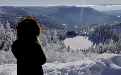 En kvinna i vinterkläder står med ryggen mot kameran och ser ner mot en sjö som är omringad av skog och berg. Allt är täckt av snö utom kvinnan.