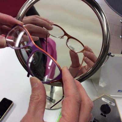 Optikko pitelee punaisia silmälaseja kädessään suurentavan peilin edessä.
