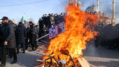 Brinnande plakat på hög. I bakgrunden mörkklädda demonstranter och längst bak till höger en moské.