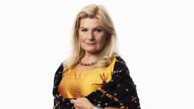 En kvinna klädd i gul silkesblus och svart sjal med färggranna streck. Hon har blont hår till axlarna, målade ögon och är i 50-årsåldern. Hon ser rakt in i kameran.