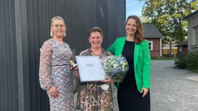 Tre kvinnor står vid en husvägg. Den mittersta visar upp en utmärkelse. De heter Henna Ljung. Hilkka Lindqvist och Petra Theman.