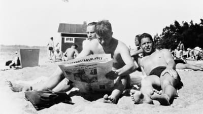 Tre män sitter på stranden. En läser tidningen. 