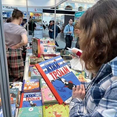 Nuori nainen tarkastelee Muumi-kuvakirjaa messukojun luona. Näyttelypöydällä on lisää Muumi-kirjoja ja muita lastenkirjoja.