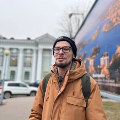 Ruskeaan hupputakkiin ja mustaan pipoon pukeutunut nuorehko mies katsoo hymyillen kameraan.