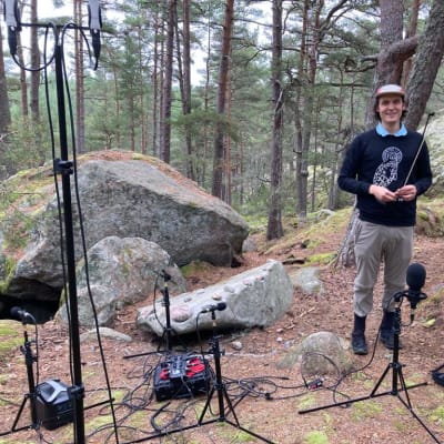 Mies seisoo metsässä suuren kiven vieressä. Etualalla on mikrofonitelineitä ja muuta äänittäm iseen liittyvää laitteistoa. 
