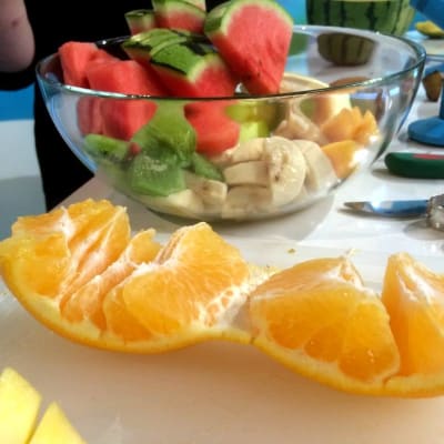 Kuorittu appelsiini laudalla ja pilkottuja hedelmiä vadissa.