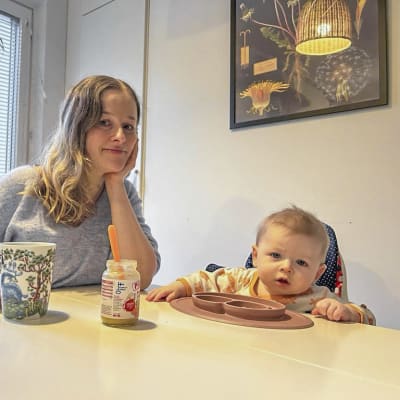 En kvinna och ett spädbarn sitter vid ett bord. De ser båda in i kameran med allvarlig min. På bordet finns mat.