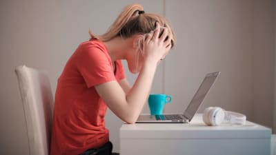 Kvinna i röd t-shirt sitter vid ett bord med uppfälld laptop och lutar huvudet i händerna 