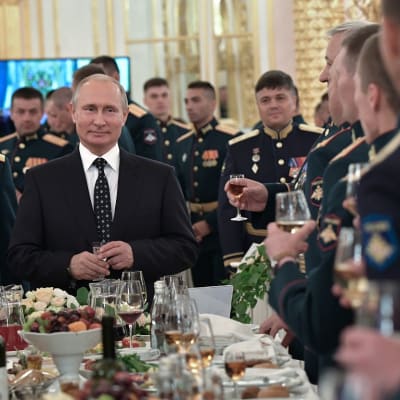 Putin seisoo sampanjalasi kädessään tummassa puvussa pöydän päässä. Pöydän ääressä ja taustalla näkyy sotilashenkilöitä juhlaunivormuissa lasit käsissään.