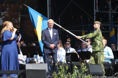 Sarah Dawn Finer talar i mikrofon medan kungen står framför en svensk flagga som hålls upp av en militär.